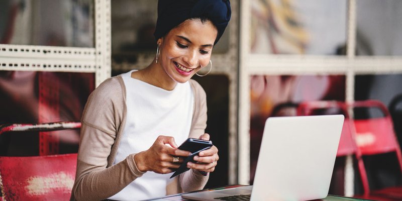 A professional woman sends a message on her phone while working in front of her laptop in a mixed outdoor workspace