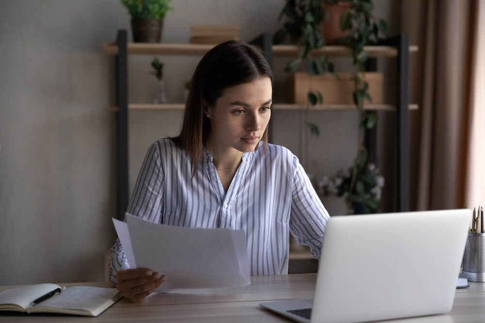 Focused busy young office employee working at laptop, reviewing online reports, checking financial documents, holding paper contracts, insurance agreement. Business woman doing paperwork job