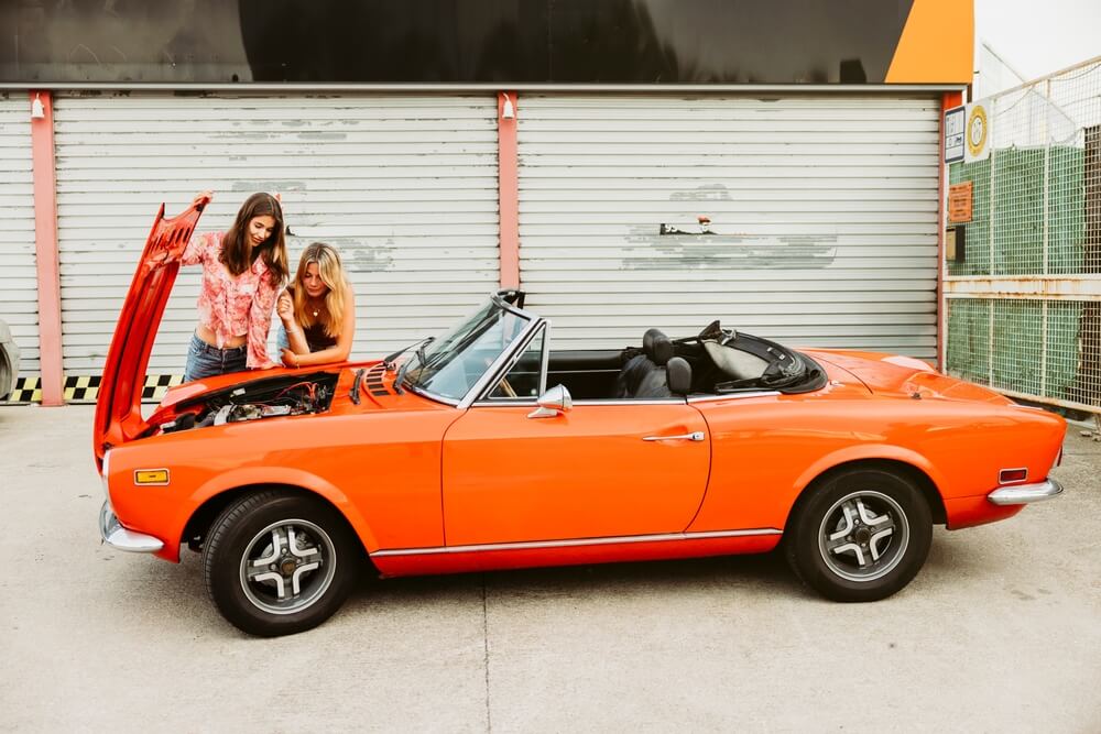 Two young women checking the engine of their vintage red cabriolet car during a road trip