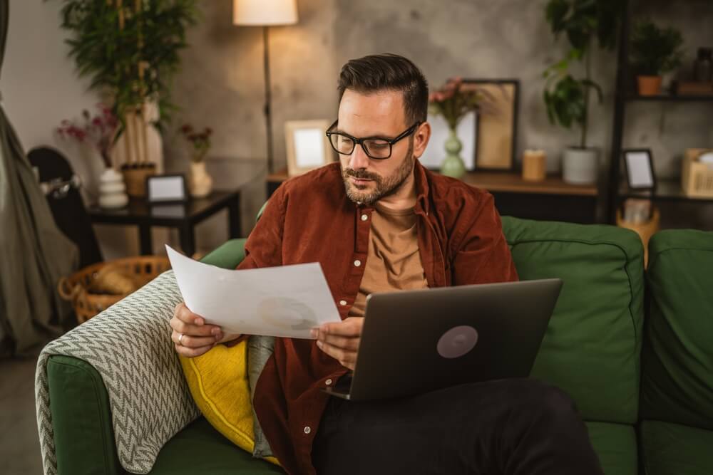 Man sitting at home and looking at laptop while hold a paper
