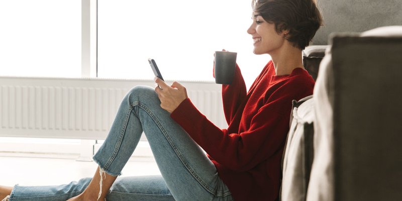 A stylish young woman with short hair sits on the floor drinking a coffee and interacting on her mobile phone